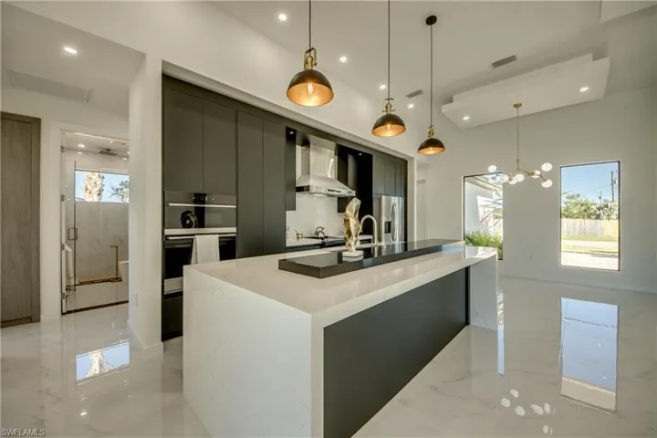 Kitchen featuring a center island with sink, wall chimney exhaust hood, stainless steel appliances, and decorative light fixtures