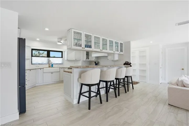White kitchen featuring a farmhouse sink, white cabinetry, a large window, a breakfast bar with seating, and wood-finish flooring - Virtually Edited Image