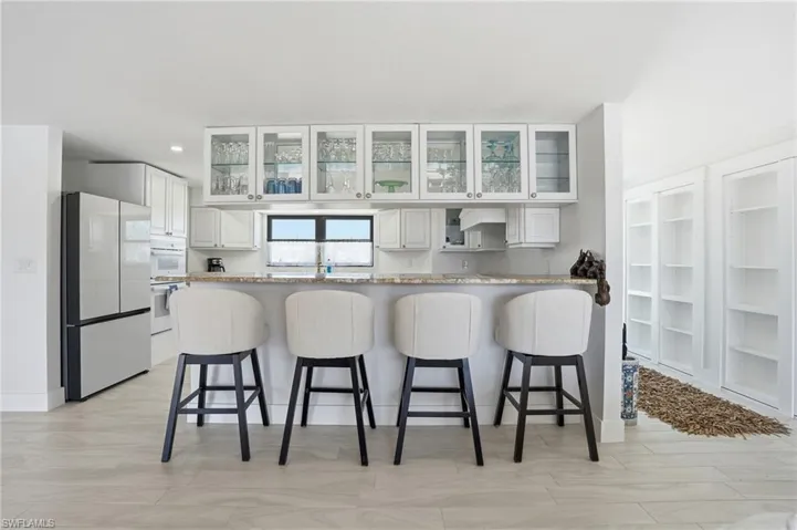 Kitchen featuring a breakfast bar with stone countertop, white cabinetry with glass-front uppers, and stainless steel appliances - Virtually Edited Image