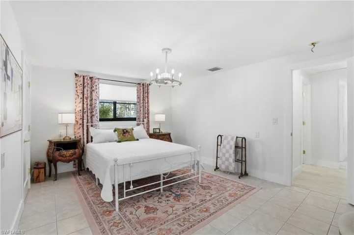 Spacious room featuring light-toned tile flooring, a large window with roller shades, and a chandelier-style ceiling light - Virtually Edited Image