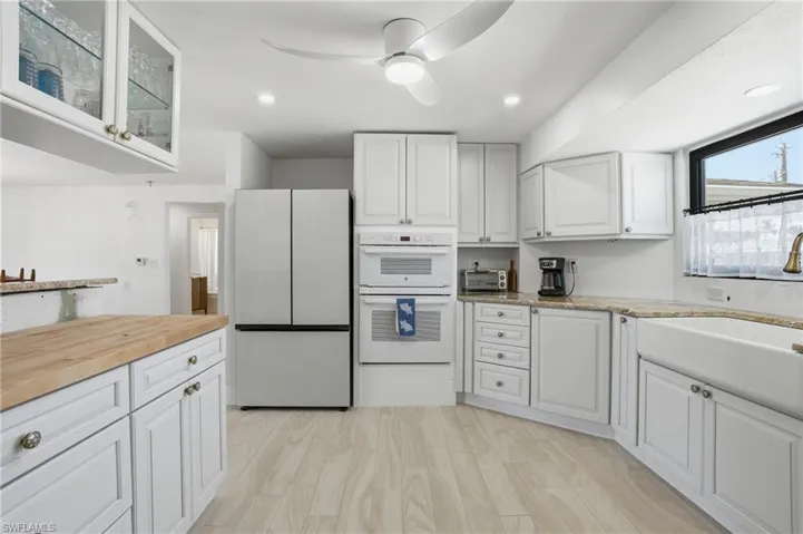 Bright kitchen featuring light wood-finish flooring, white cabinetry with round hardware, a modern white refrigerator, a double wall oven, and light granite-finish countertops - Virtually Edited Image