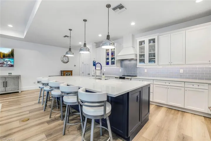 Kitchen featuring decorative light fixtures, custom exhaust hood, decorative backsplash, light wood-type flooring, and a center island with sink