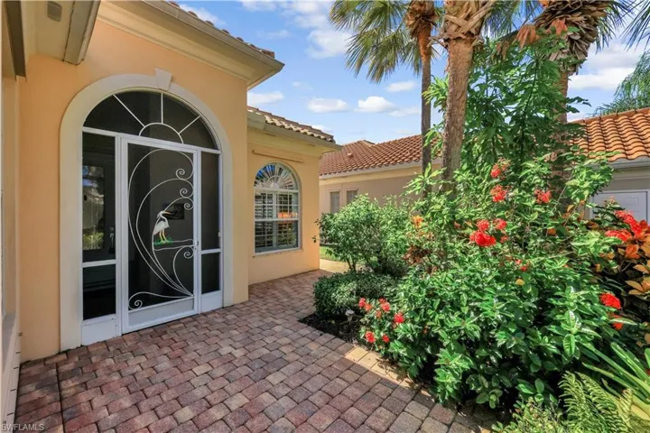 Stucco exterior entry featuring a decorative screened door with arch transom, terracotta-toned tile roofing, and a paved brick walkway surrounded by mature tropical landscaping