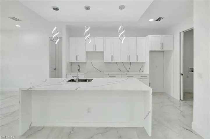 Kitchen with light stone countertops, a center island with sink, decorative light fixtures, and white cabinets