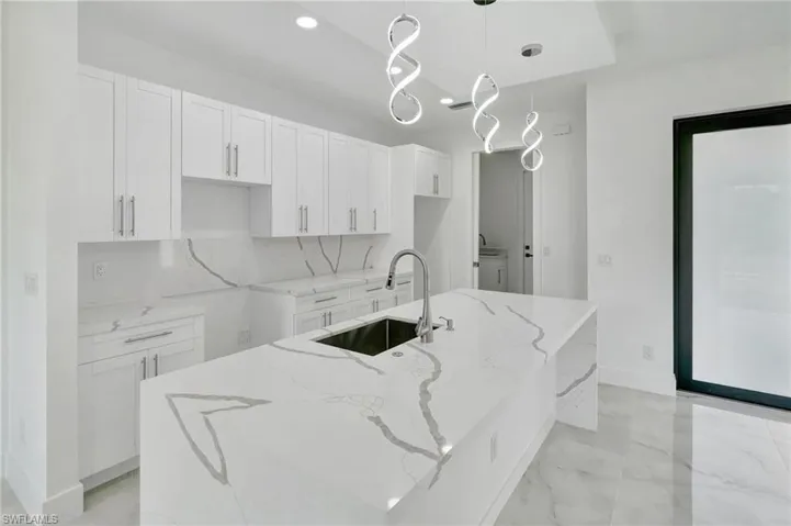 Kitchen featuring light stone counters, a center island with sink, white cabinetry, and decorative light fixtures