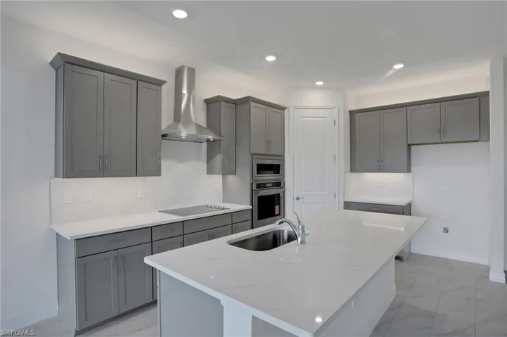 Kitchen featuring light stone countertops, an island with sink, gray cabinetry, recessed lighting, and stainless steel appliances