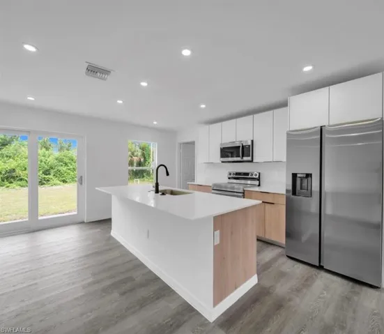 Kitchen with stainless steel appliances, a center island with sink, white cabinetry, recessed lighting, and light brown cabinets