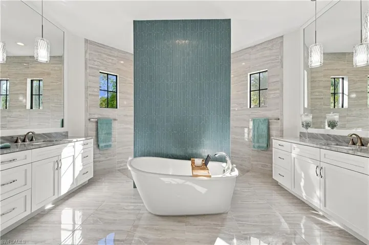 Freestanding soaking tub set against a blue decorative tile accent wall, flanked by dual white cabinetry vanities with stone countertops, large mirrors, and pendant lighting