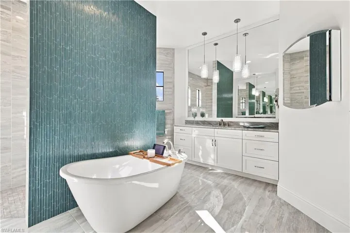Freestanding soaking tub set against a blue glass mosaic tile accent wall, featuring a white dual-sink vanity, stone-finish countertops, full-width wall mirror, and three glass pendant lights