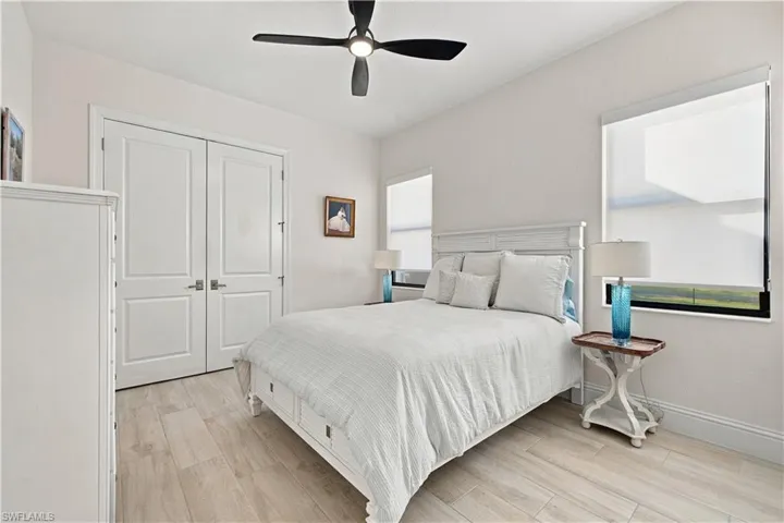 Light-toned wood-finish tile flooring, matte black ceiling fan, dual-panel closet doors, and multiple windows with roller shades