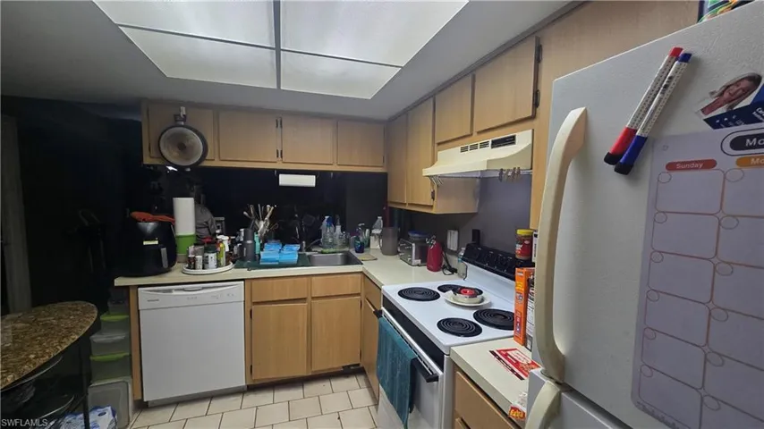 Kitchen featuring white appliances, light countertops, under cabinet range hood, light tile patterned floors, and brown cabinets