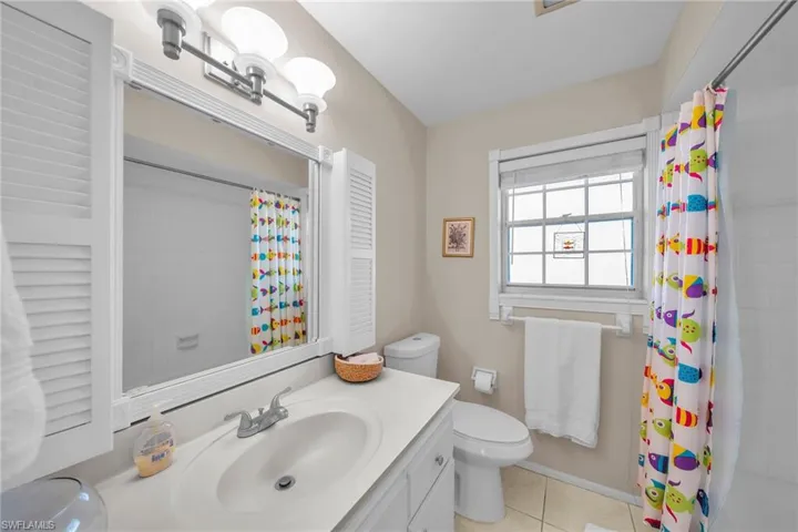 Bathroom featuring a single vanity with an integrated sink, a framed mirror with louvered side panels, overhead bar lighting, a window with blinds, and tiled flooring