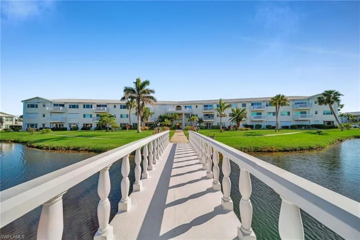 Waterfront multi-story building featuring individual balconies, surrounding water features, a white balustrade bridge, and manicured green spaces with mature palm trees