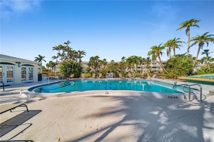 Outdoor swimming pool with integrated steps, surrounded by a concrete deck, mature palm trees, and a low-rise building featuring double French doors