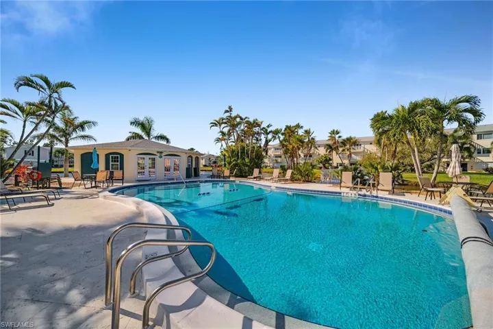Community swimming pool with a surrounding paved deck, palm trees, and a pool house featuring double doors and an awning