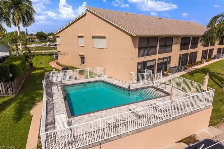 Fenced outdoor swimming pool with a paver deck, surrounded by mature landscaping and a multi-story building featuring screened balconies