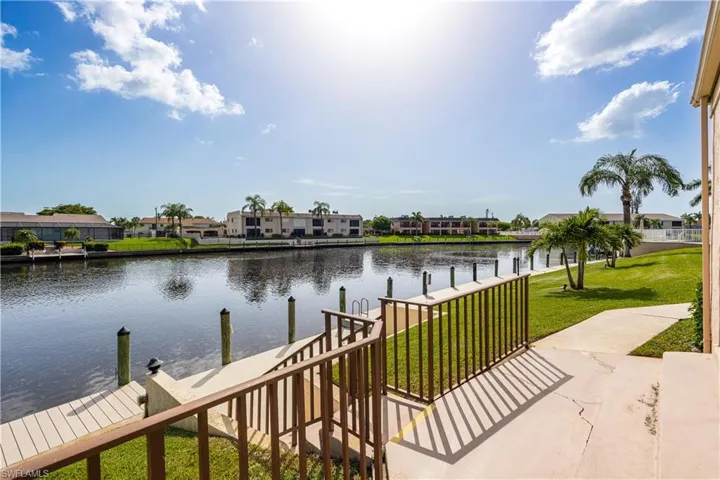 Waterfront property featuring a private dock with pilings, a concrete walkway, mature palm trees, and a manicured lawn