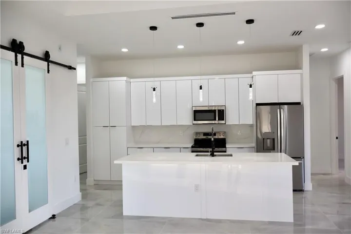 Kitchen featuring stainless steel appliances, white cabinets, decorative light fixtures, a barn door, and a kitchen island with sink