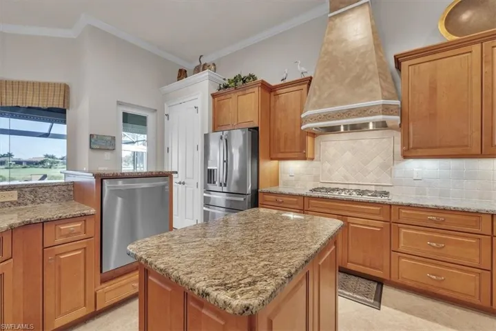Kitchen with custom range hood, brown cabinetry, appliances with stainless steel finishes, light stone counters, and crown molding