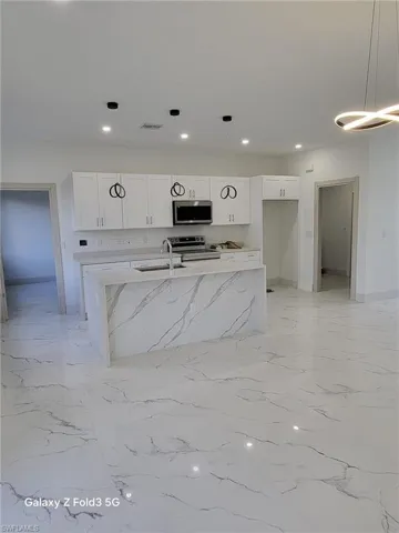 Kitchen with hanging light fixtures, white cabinets, light stone countertops, stainless steel appliances, and a kitchen island with sink