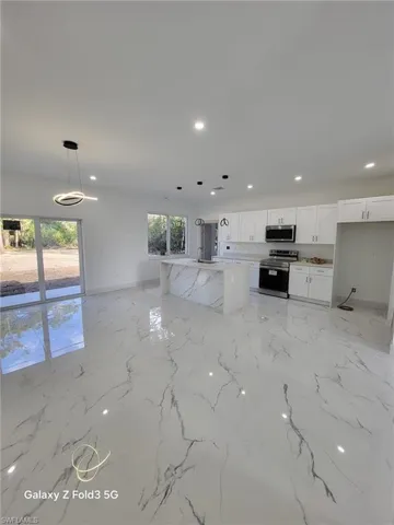 Kitchen with white cabinets, stainless steel appliances, an island with sink, decorative light fixtures, and light stone counters