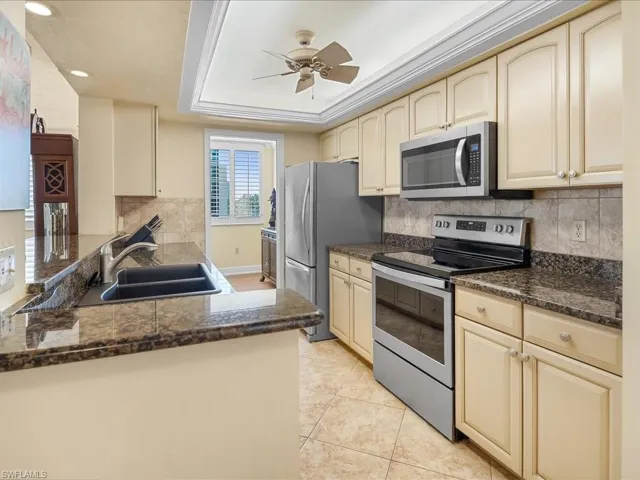 Kitchen featuring stainless steel appliances, granite countertops, cream cabinets, lighted tray ceiling, backsplash, a tray ceiling, tile floor and a ceiling fan