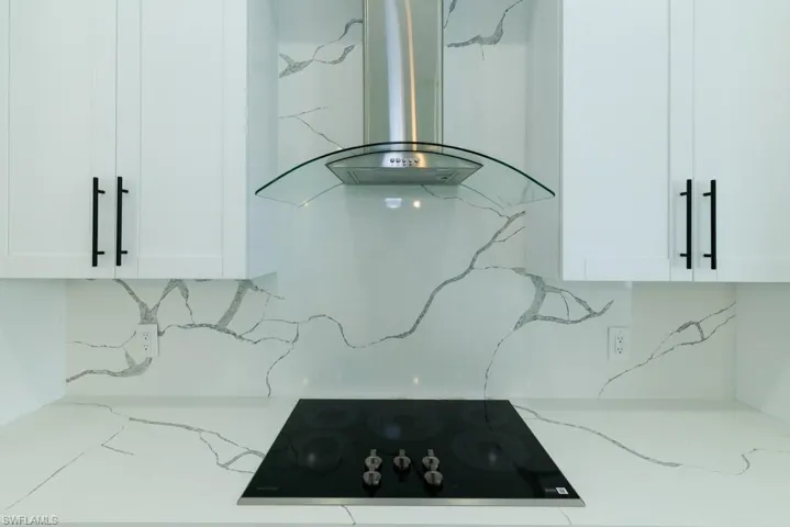 Kitchen view of range hood, black electric stovetop, light stone counters, and backsplash
