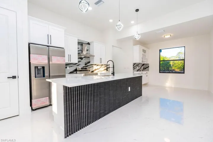 Kitchen with stainless steel fridge, white cabinets, hanging light fixtures, light stone counters, and a center island with sink