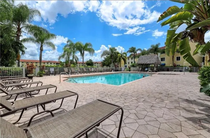 Resort-style pool area featuring a rectangular swimming pool, extensive paver deck, multiple lounge chairs, lush palm trees, and a thatched-roof cabana