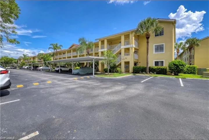 Multi-story residential complex featuring a pale yellow exterior, a paved parking area, and carports