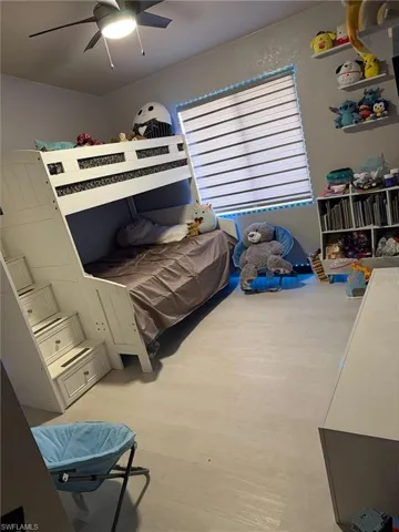 White bunk bed with built-in stairway storage, light wood-finish flooring, a ceiling fan with integrated lighting, a window with horizontal blinds, and wall-mounted shelving