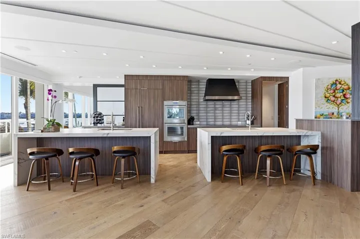 Two tone kitchen with a breakfast bar area, light stone counters, modern cabinets, double oven, and recessed lighting