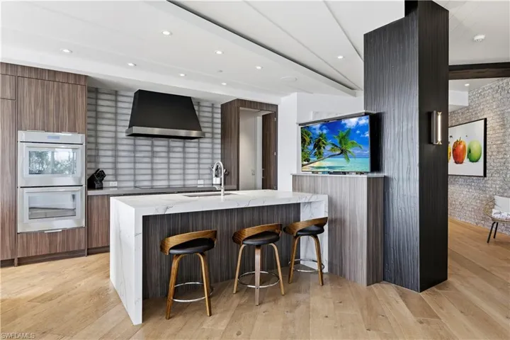 Dual tone kitchen with light stone counters, a breakfast bar area, a kitchen island with sink, stainless steel double oven, and modern cabinets