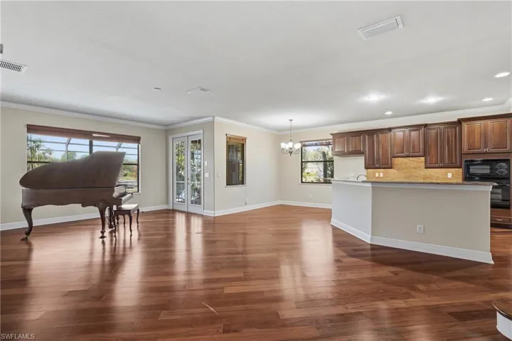 Open-concept floor plan showcasing a spacious family room with piano, casual dining beneath an elegant chandelier, and French doors opening to the pool area with convenient access to a full cabana bath.