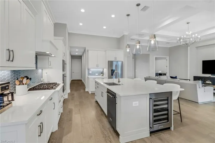 Kitchen featuring white cabinets, gas cooktop, stainless steel appliances. Large island with sink, hanging pendant lights, dishwasher and wine cooler
