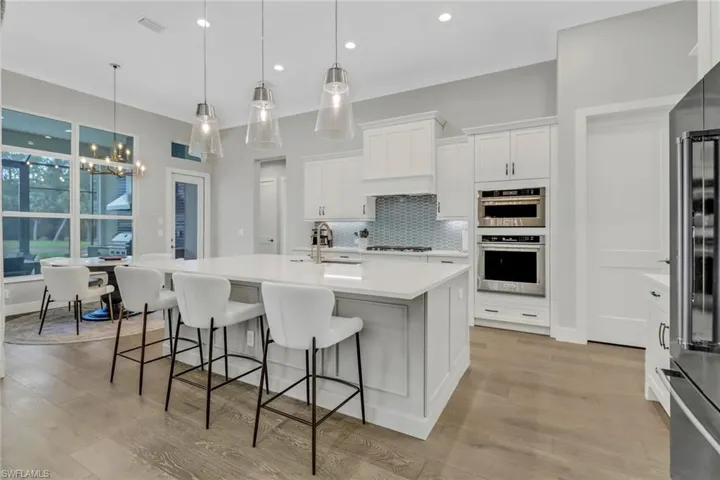 Kitchen with white cabinetry, hanging pendant lights, light hardwood floors and a kitchen island with sink and wine cooler.