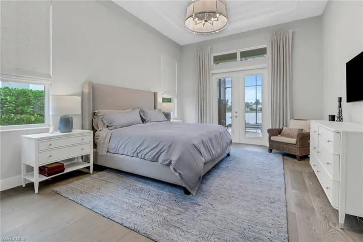 Primary bedroom featuring french doors leading to the lanai, light hardwood floors, a notable chandelier, custom tray ceiling and custom drapes.
