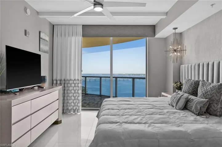 Bedroom featuring a water view, light tile patterned flooring, ceiling fan with notable chandelier, beam ceiling, and access to outside