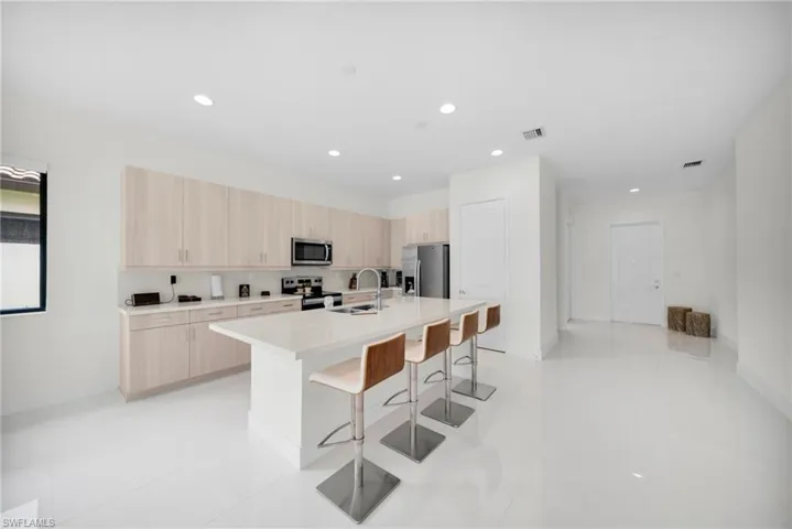 Spacious kitchen featuring light wood-finish cabinetry, stainless steel appliances, a large island with countertop seating, recessed lighting, and white tile flooring