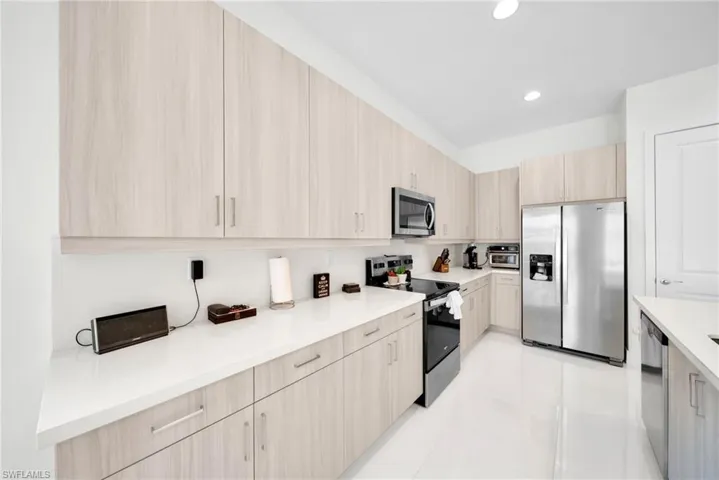 Contemporary kitchen featuring extensive light wood-finish cabinetry, bright white countertops, and stainless steel appliances including a side-by-side refrigerator, microwave, and dishwasher