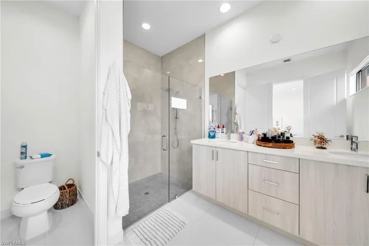Spacious bathroom featuring a glass-enclosed shower with tile surround, a dual vanity with a light wood-finish cabinet and white countertop, and recessed ceiling lighting