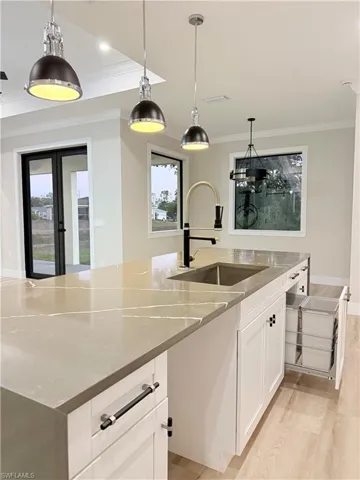 Kitchen featuring decorative light fixtures, crown molding, white cabinets, light stone countertops, and a kitchen island with sink