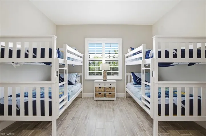 Spacious room featuring two sets of white bunk beds, light wood-finish flooring, and a central window with white plantation shutters