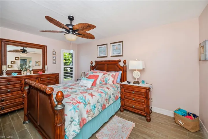 Bedroom featuring wood-look tile flooring, a detailed ceiling fan with leaf-shaped blades, and a window with white louvered shutters