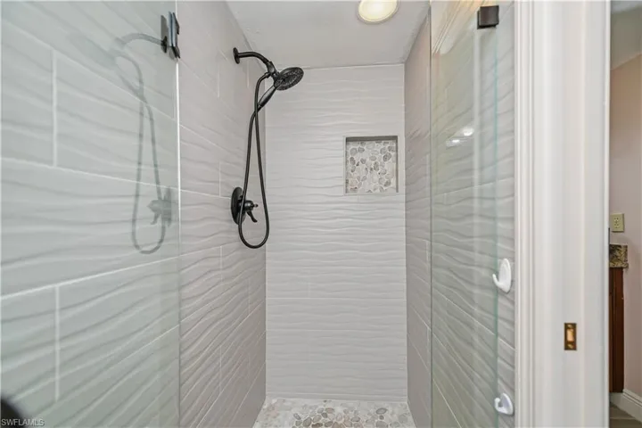 Walk-in shower featuring textured wave-patterned wall tiles, pebble-style floor tiling, matte black shower fixtures, a built-in shower niche with mosaic stone accents, and clear glass shower doors