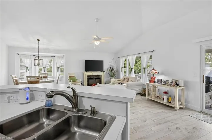 Kitchen bar with a double basin stainless steel sink, extending to a living area featuring vaulted ceilings, light wood-finish flooring, multiple windows, and a sliding glass door