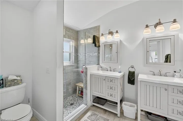 Bathroom featuring a glass-enclosed shower with pebble flooring and a window, dual vanity sinks with white cabinetry, and individual framed mirrors