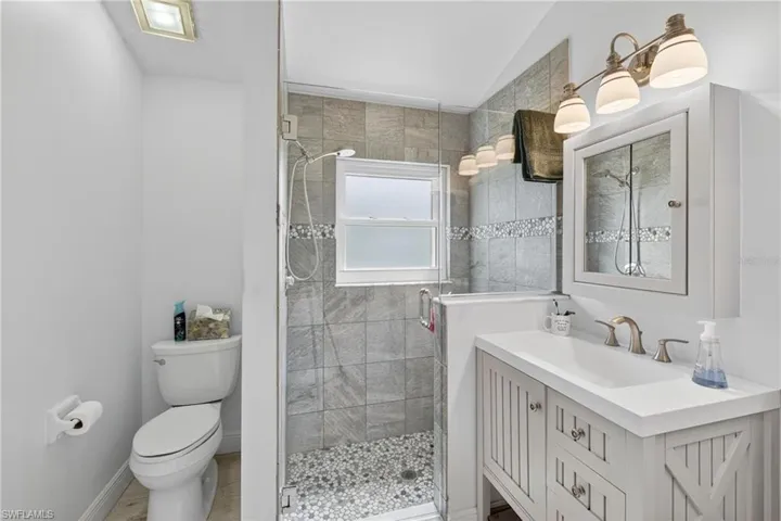 Bathroom featuring a standing shower with clear glass enclosure and pebble floor, gray tile shower surround with decorative mosaic accent, white vanity with integrated sink, brushed nickel faucet, and a wall-mounted medicine cabinet with mirrored door