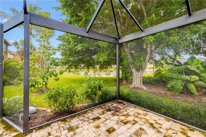 Screened outdoor living area with paver flooring, overlooking a manicured lawn and mature trees, with a water feature beyond