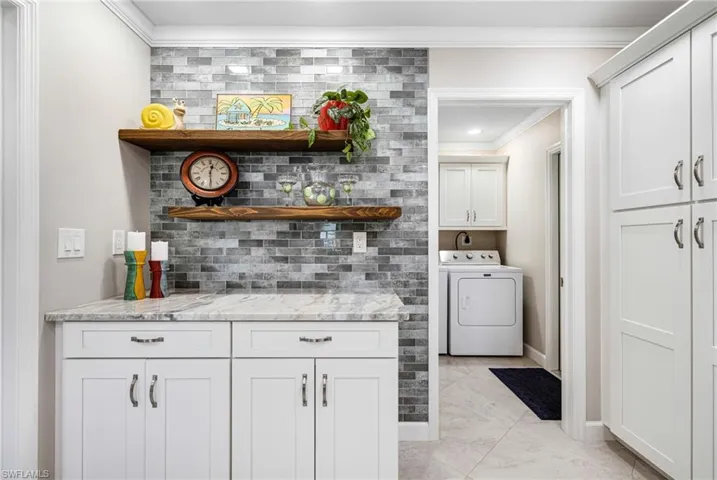 Bar with light stone counters, washer and clothes dryer, crown molding, backsplash, and white cabinetry